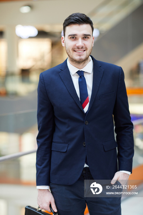 Smiling handsome young male bank manager in suit standing in business hall and looking at camera while holding hand in pocket