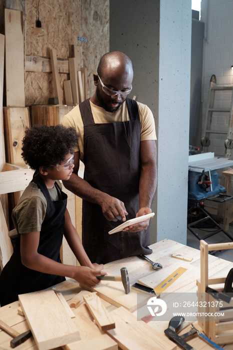 Vertical portrait of African-American father teaching son carpentry while working together in woodworking shop