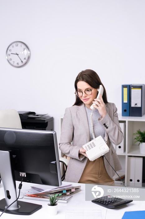 Content attractive young businesswoman in glasses standing at desk and talking on phone in office