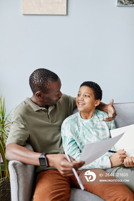 Vertical portrait of smiling African American girl doing homework with father while sitting on sofa