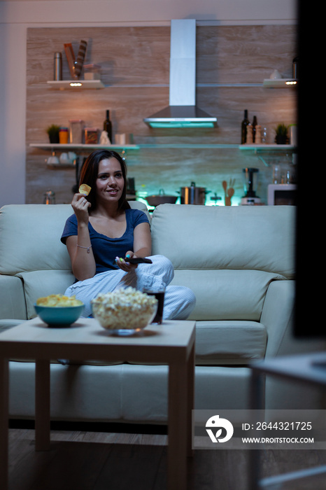 Woman enjoying the evening watching tv series at home sitting on comfortable couch dressed in pajamas. Excited amused home alone lady eating snacks and drinking juice on cozy sofa in living room.