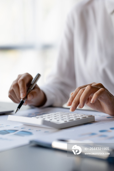 Businessman is using a calculator to calculate company financial figures from earnings papers, a businessman sitting in his office where the company financial chart is placed.