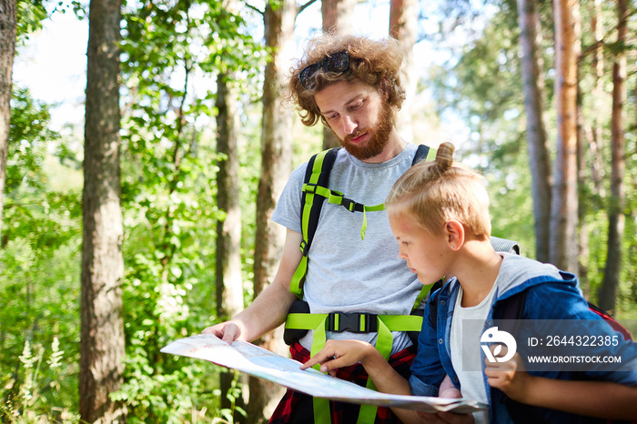 One of boy scouts with backpacks pointing at map while looking for dwelling area in the forest