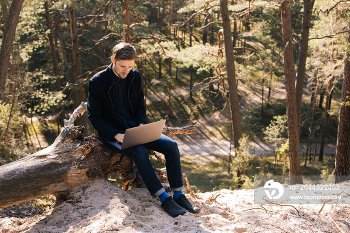 Man using a laptop in the forest. Young man sitting on an old tree strain work remotely in nature. Work everywhere traveler blogger outdoors. Businessman sitting in forest Remote working concept