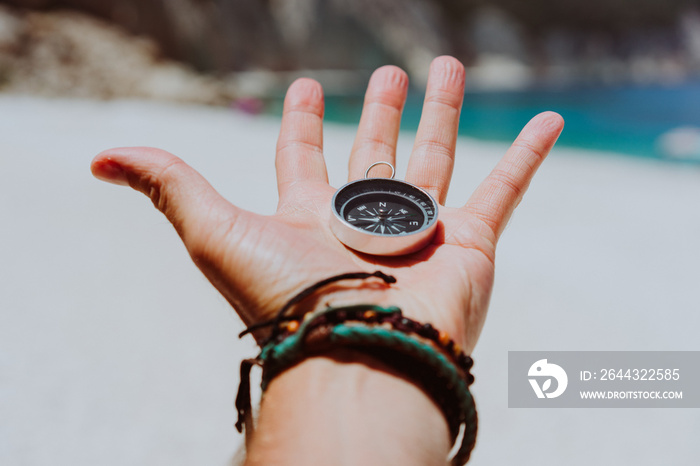 Open palm with stretched fingers holding black metal compass against white sandy beach. Find your way or goal concept. Point of view pov