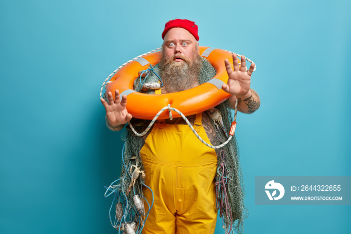 Scared bearded fiisherman afraids of swimming, raises palms in protective gesture, scared of drowning in sea, poses with fishing gears, isolated on blue background. Frightened bearded male angler