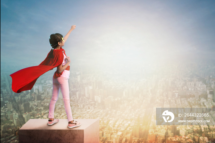 Super girl child show power on rooftop of building ,little kid wearing red hero cloak show hand up to sky and city in sun set, active dream and motivation idea concept