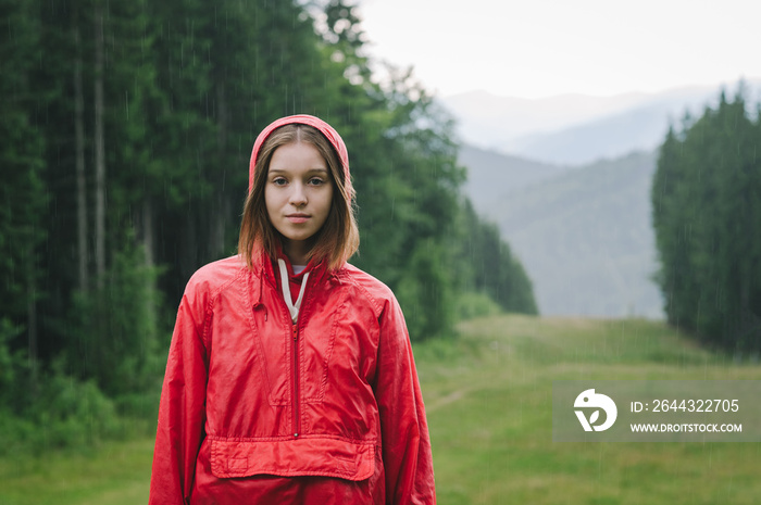 Portrait of a young beautiful lady standing on the mountain hill in the pouring rain wearing a red waterproof jacket, feeling peaceful and calm.