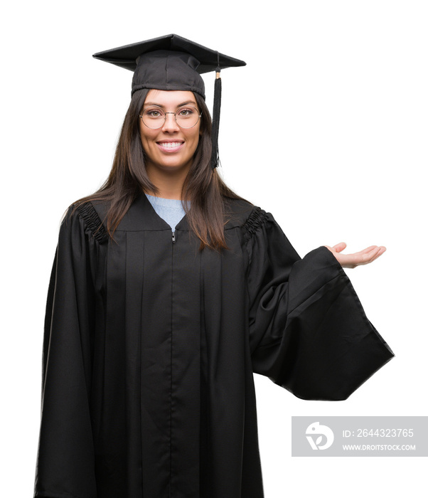 Young hispanic woman wearing graduated cap and uniform smiling cheerful presenting and pointing with palm of hand looking at the camera.