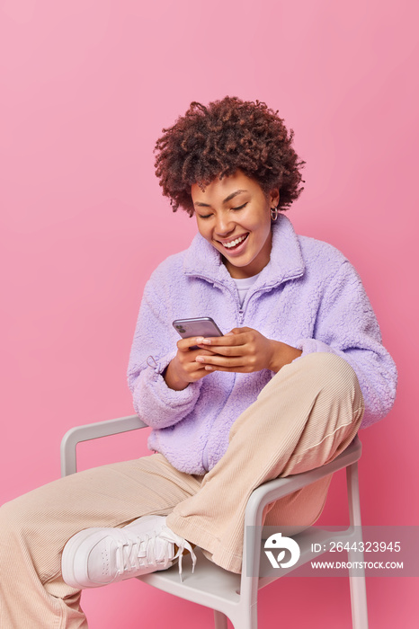 Happy glad relaxed woman uses mobile phone updates information on website uses free internet connection dressed in stylish clothes sits on chair isolated over pink background sends text messages