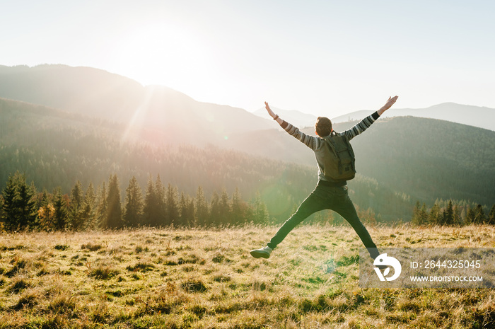 Happy traveler man with backpack jump on sunset in autumn day with open hands. Back view. Hipster enjoying sunrise on peak mountains. Tourist travel concept. Hiker looking sunlight in country Europa.