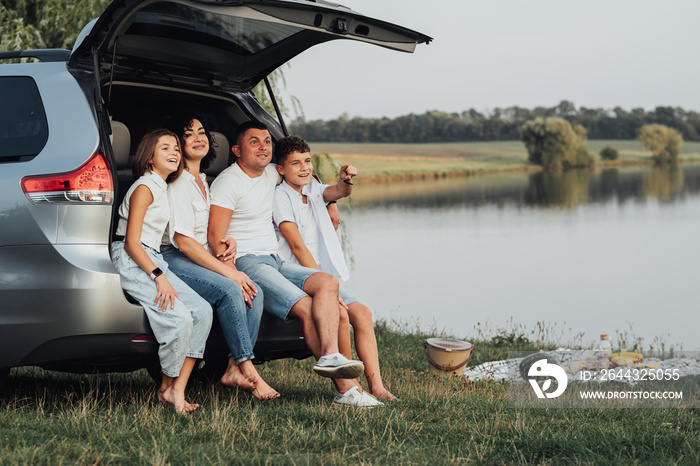 Happy Four Caucasian Members Family Sitting in Trunk of Minivan Car, Mother and Father with Two Teenage Children, Son and Daughter Having Weekend Picnic Outdoors by the Lake