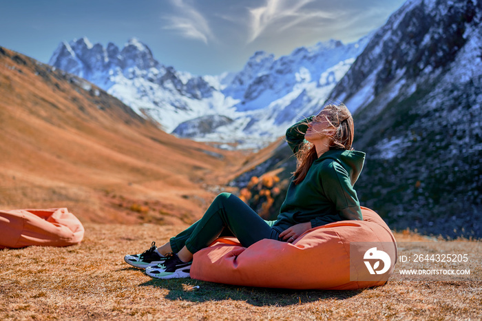 Wanderer girl sitting on soft chair during resting in a camping. Traveler enjoying scenic landscape while traveling in a mountain valley