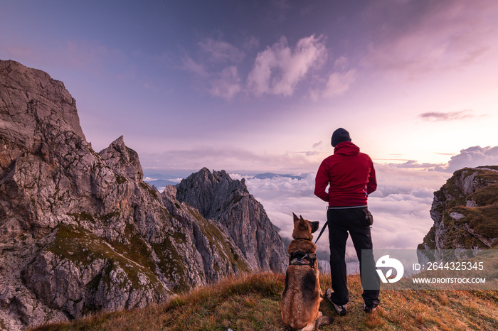 Adventure Man with Dog at High Mountains Peak at Sunrise. Togetherness and Friendship Concept