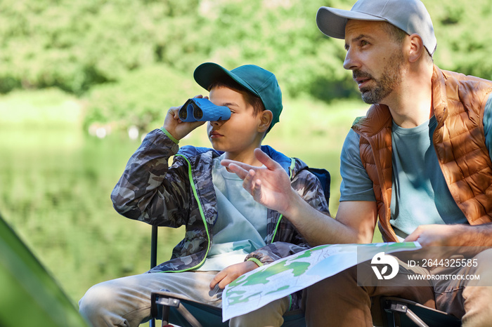 Portrait of teenage boy looking in binoculars while enjoying camping trip with father, copy space