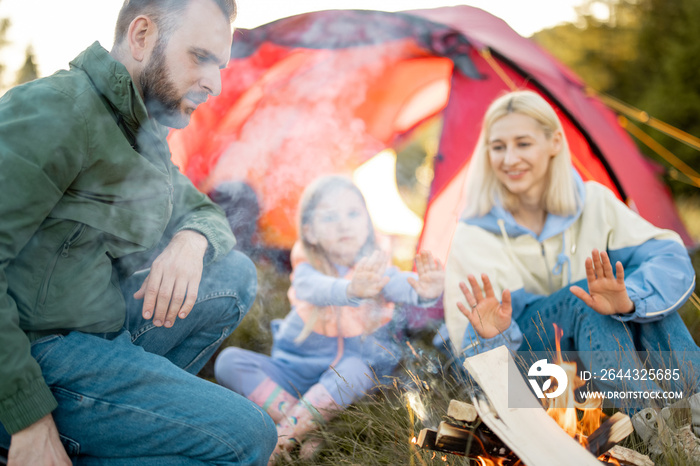 Young adult couple sit with a little girl and have fun by the campfire, traveling with tent on nature. Family spend summer time hiking in the mountains