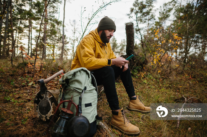 Survival in the wild. A bearded man is resting on a fallen tree in the middle of a forest.