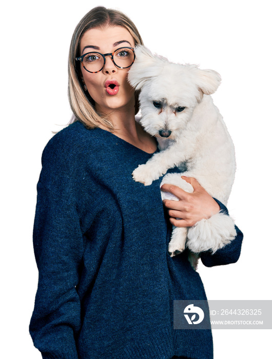 Young blonde woman hugging cute dog scared and amazed with open mouth for surprise, disbelief face