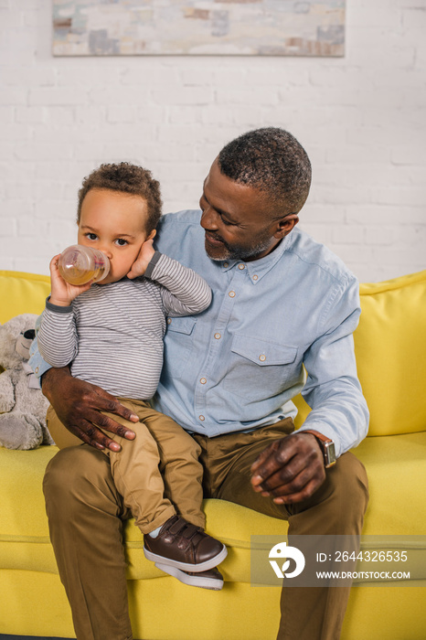 happy african american grandfather looking at little grandson drinking from baby bottle at home