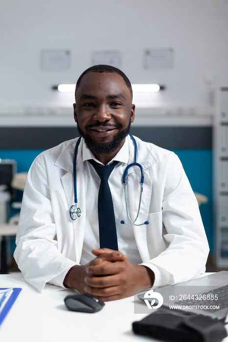 Portrait of african american therapist doctor sitting at desk in hospital office working at medical treatment. Practitioner man analyzing sickness symptoms expertise. Healthcare service