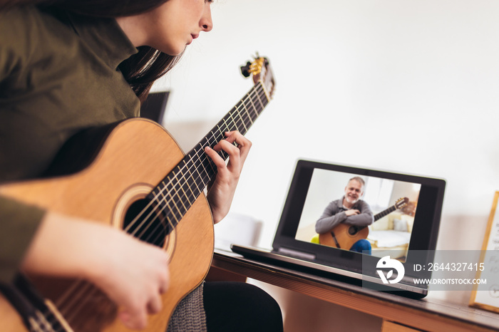 Focused girl playing acoustic guitar and watching online course on laptop while practicing at home. Online training, online classes.