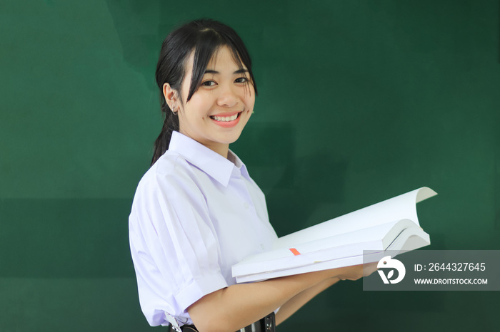 Smiling happy young portrait pretty Asian student girl holding book at green board in high school or college uniform, smile at front classroom for learning. Education learning in college