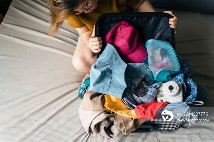 Asian woman looking at her messy belongings such as sunglasses, polaroid camera, cosmetic bag and small bag, try to fit everything in the luggage with struggling. Packing to go on a getaway vacation.