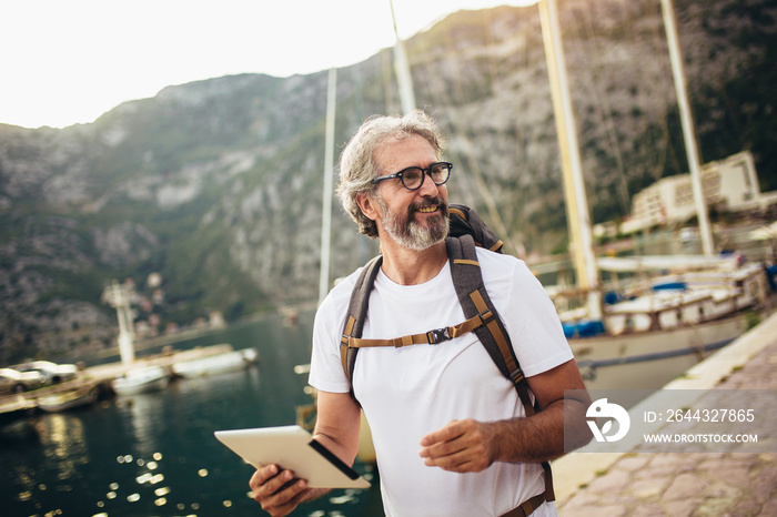 Smiling tourist mature man standing with digital tablet and backpack near the sea.