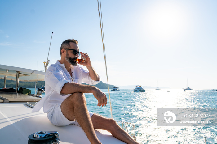 Caucasian man enjoy outdoor luxury lifestyle with alcoholic drinks while catamaran boat sailing at summer sunset. Handsome male relaxing outdoor leisure activity with tropical travel vacation trip