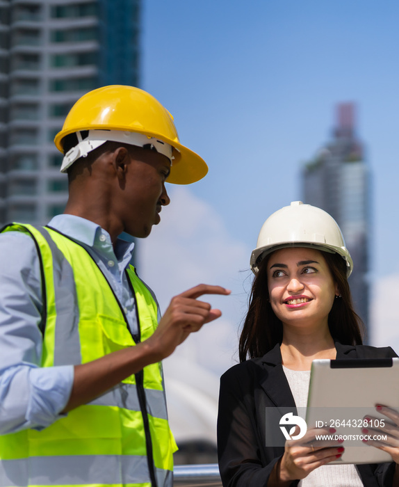 caucasian businesswoman with helmet in black suit happy working with tablet and looking at black engineer colleague at construction site outside building in city