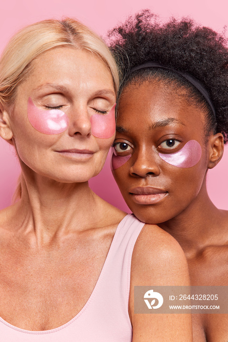 Two diverse women of different age stand closely to each other apply beauty patches under eyes remove dark circles take care of skin undergo anti aging procedures enjoy spa day at home pose indoor