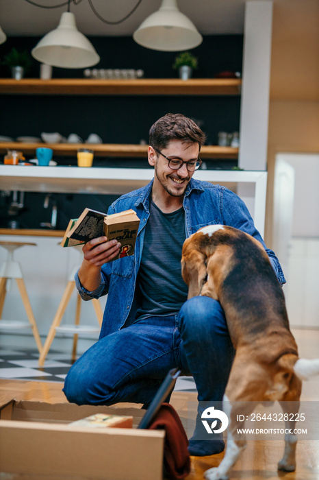 A young student man moving alone into a new apartment, taking out a book from the box while his dog is playing around.