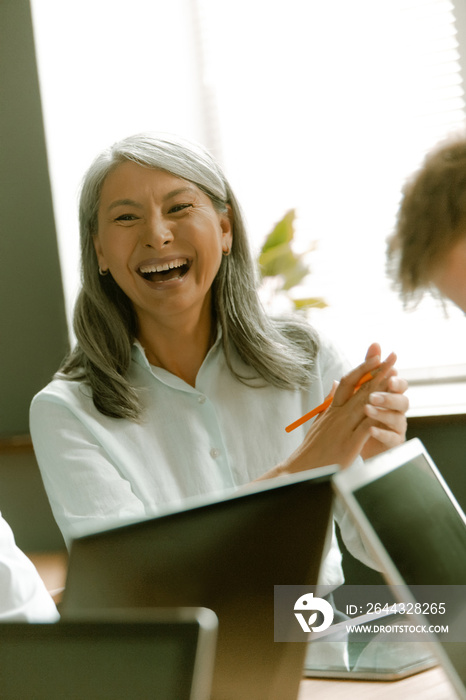 Funny Asian senior woman laughs at business meeting sitting at office desk with laptop computers on it against of window on background.