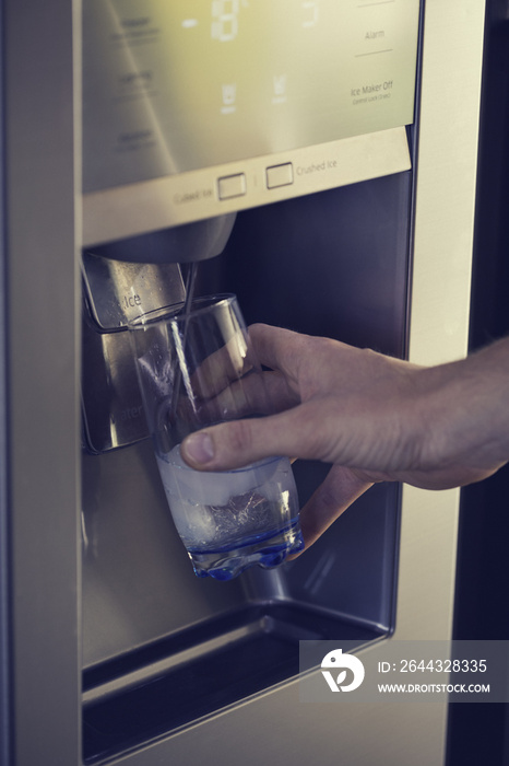 Male hand is pouring cold water and ice cubes from dispenser of home fridge.