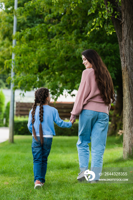 back view of mother and daughter in jeans holding hands while walking in park