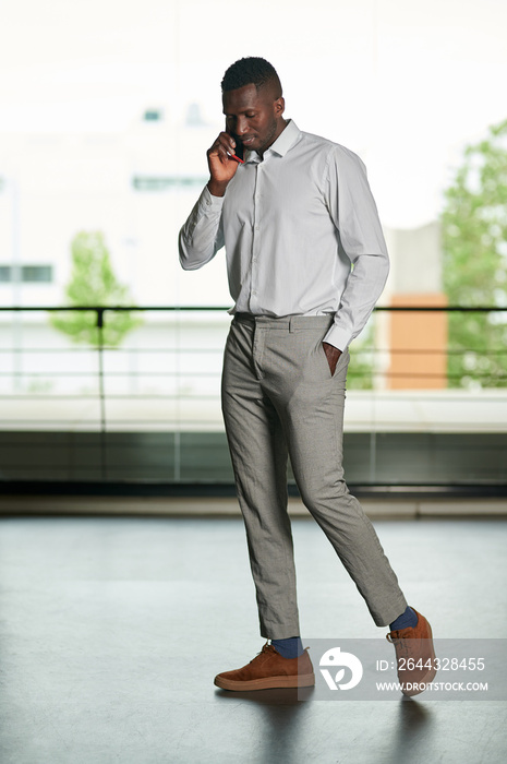 Full body of African male entrepreneur in formal wear looking away and having phone conversation while walking in corridor of office building