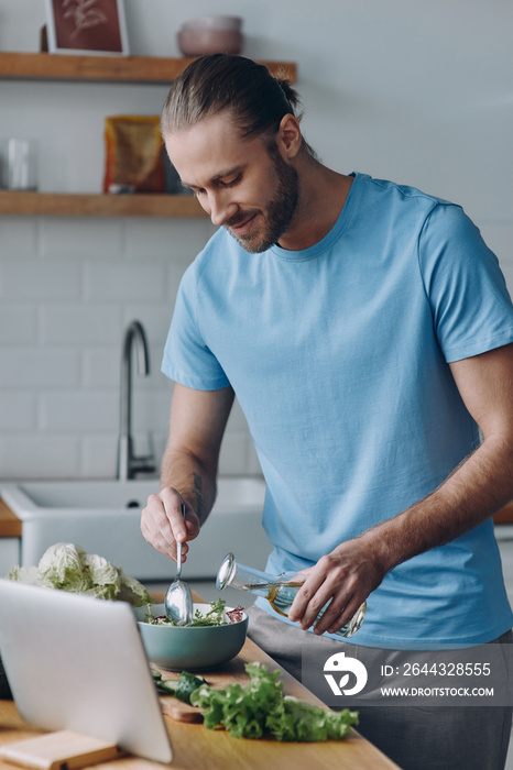 Confident young man pouring some olive oil on salad while preparing food at the kitchen