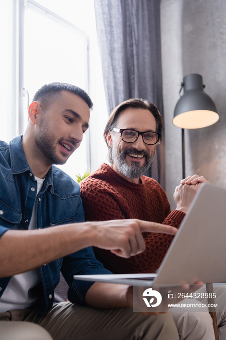 Smiling Hispanic man pointing at laptop near cheerful father, blurred foreground