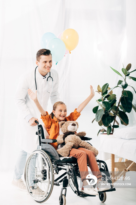 smiling pediatrist and kid with teddy bear on wheelchair in hospital