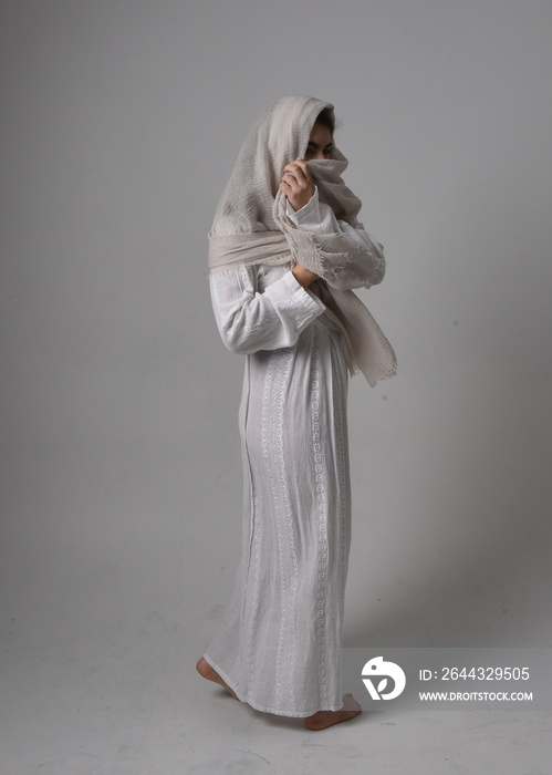 Full length portrait of young woman wearing classical white gown and a head covering veil in biblical style, sitting pose on light studio background.