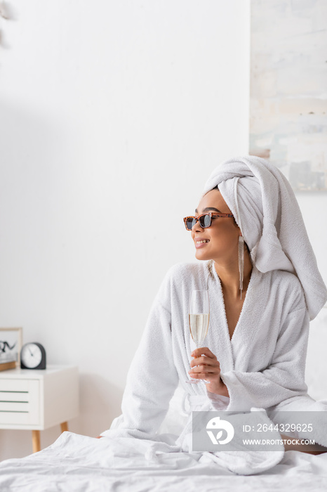smiling african american woman in terry bathrobe and trendy sunglasses holding champagne and looking away in bedroom.
