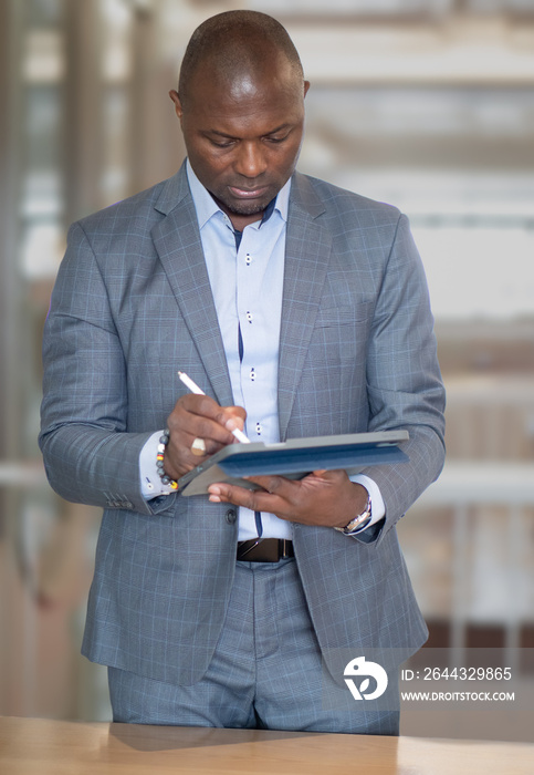 Black businessman in suit standing in modern office using tablet computer for remote working, business meeting, portrait. Handsome black manager,  executive white collar worker in diversity corporate.