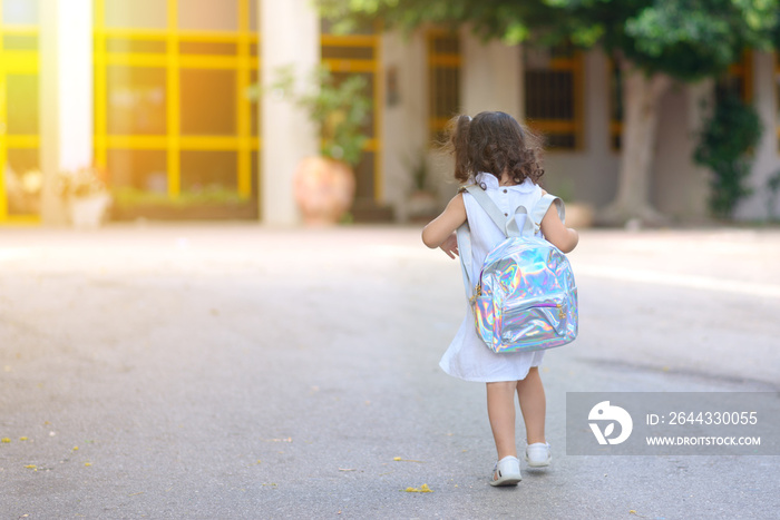 Curly cute little toddler girl back to school with holographic schoolbag or satchel, child walking to school bus. Toddler kid first day at school or preschool.