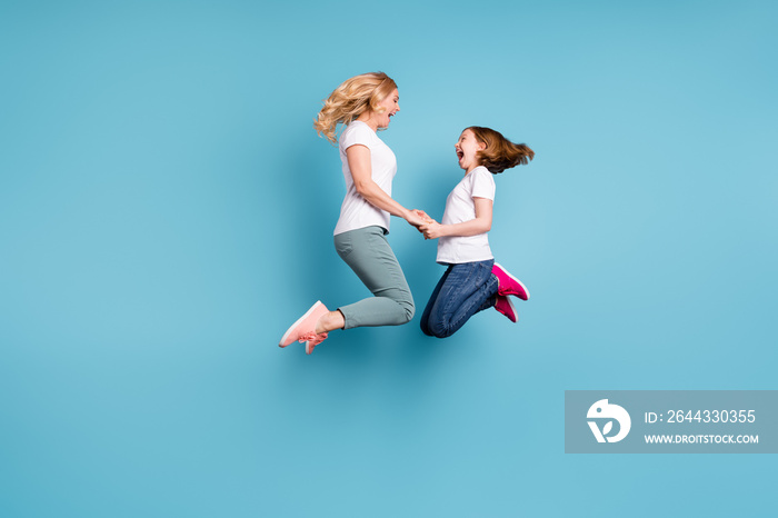 Full length profile photo of funny mom lady little daughter spend time together jumping high up hold hands rejoicing wear casual white s-shirts isolated blue color background