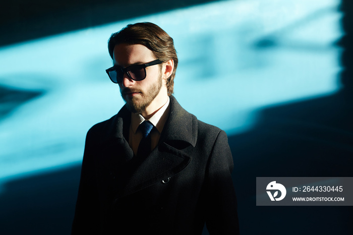 Portrait of handsome man wearing sunglasses and black coat in bright sunlight against blue background, looking away from camera