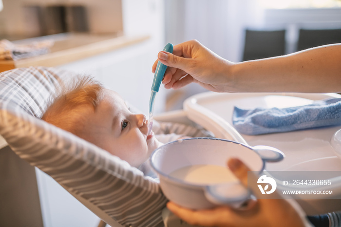 Side view of hungry little baby boy with bib sitting in his chair and taking spoon from his mother. What a delicious food.