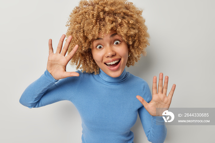 Happy cheerful curly haired European woman has surprised expression smiles broadly keeps palms raised entertains someone dressed in casual blue turtleneck poses against grey studio background