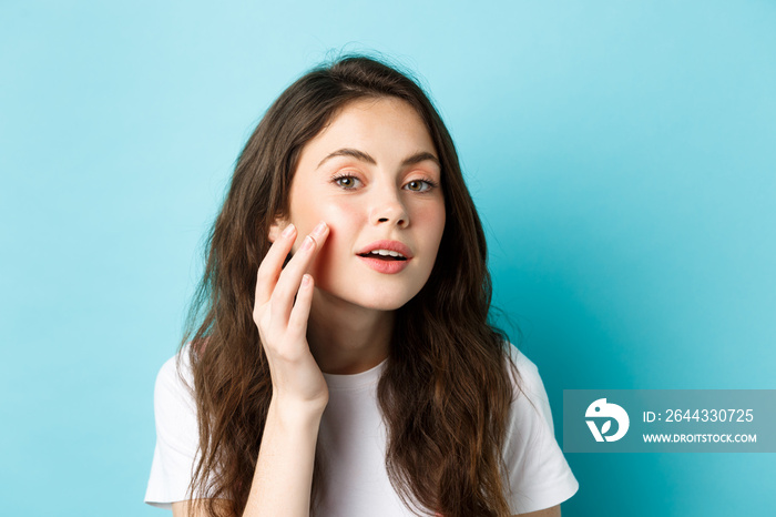 Skin care. Close up of young woman looking in mirror and touching clean glowing facial skin, checking out cosmetics after effect, apply beauty product, standing over blue background