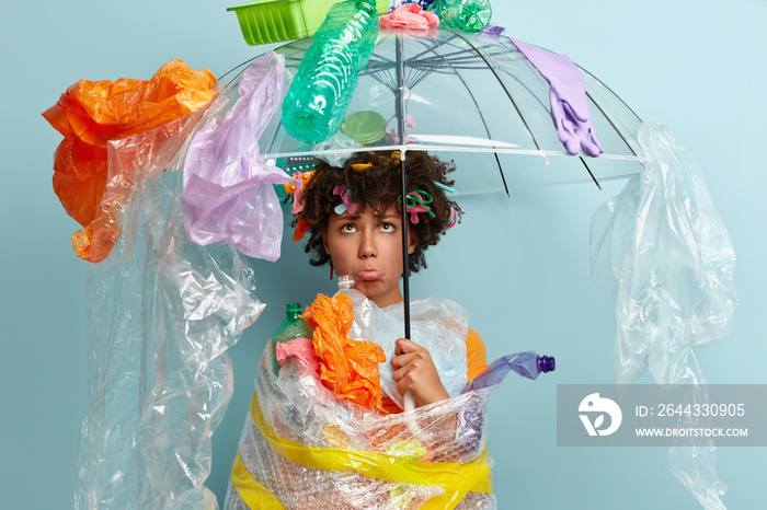 Indoor shot of depressed Afro American woman purses lower lip, being in bad mood, holds polythene umbrella with plastic garbage on top, fights against garbage, isolated against blue background