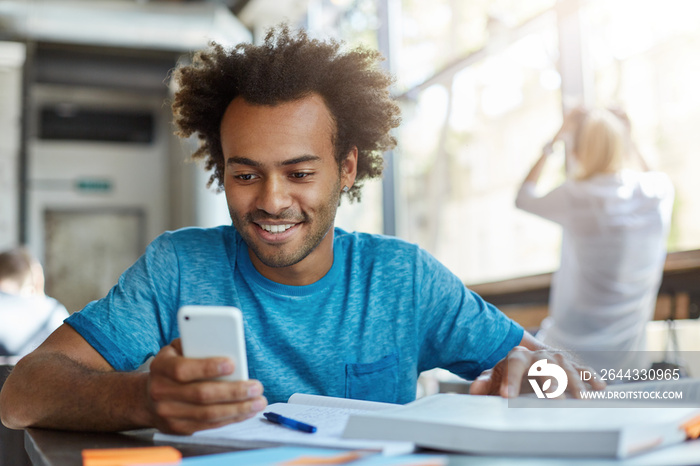 Handsome joyful student weaing blue t-shirt making video call using free onlipe app on his web-enabled smart phone, looking at screen with broad smile, sitting at cafe table with textbook and copybook